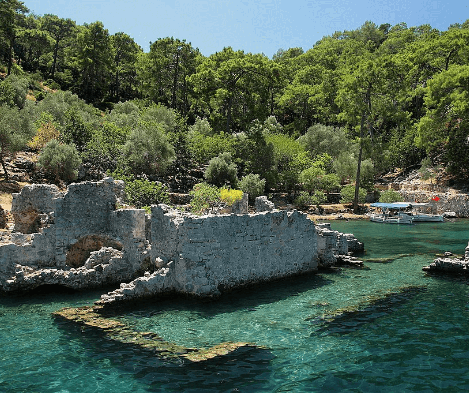 Segeltörn Türkei - Cleopatra Bucht, Ruinen von einem Steingebäudem Boote auf dem smaragdfarbenen Wasser, Bäume im Hintergrund
