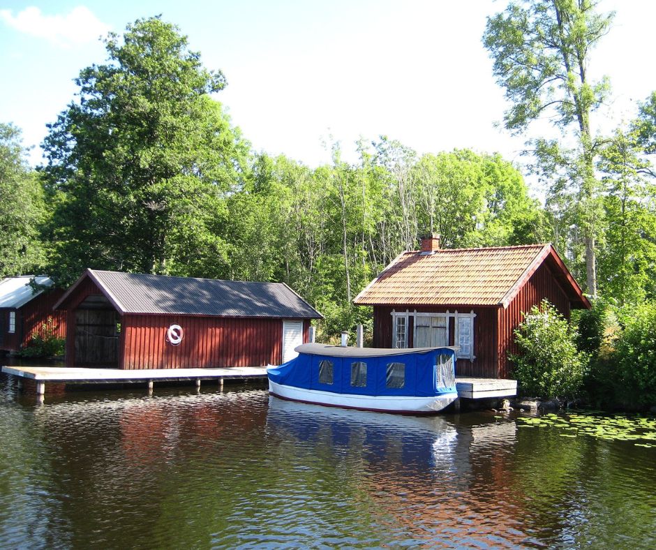 Drei schwedisch rote Häuser am Götakanal. Bei einem führt ein Steg in das Wasser. Man sieht ein Boot vor einem Haus.