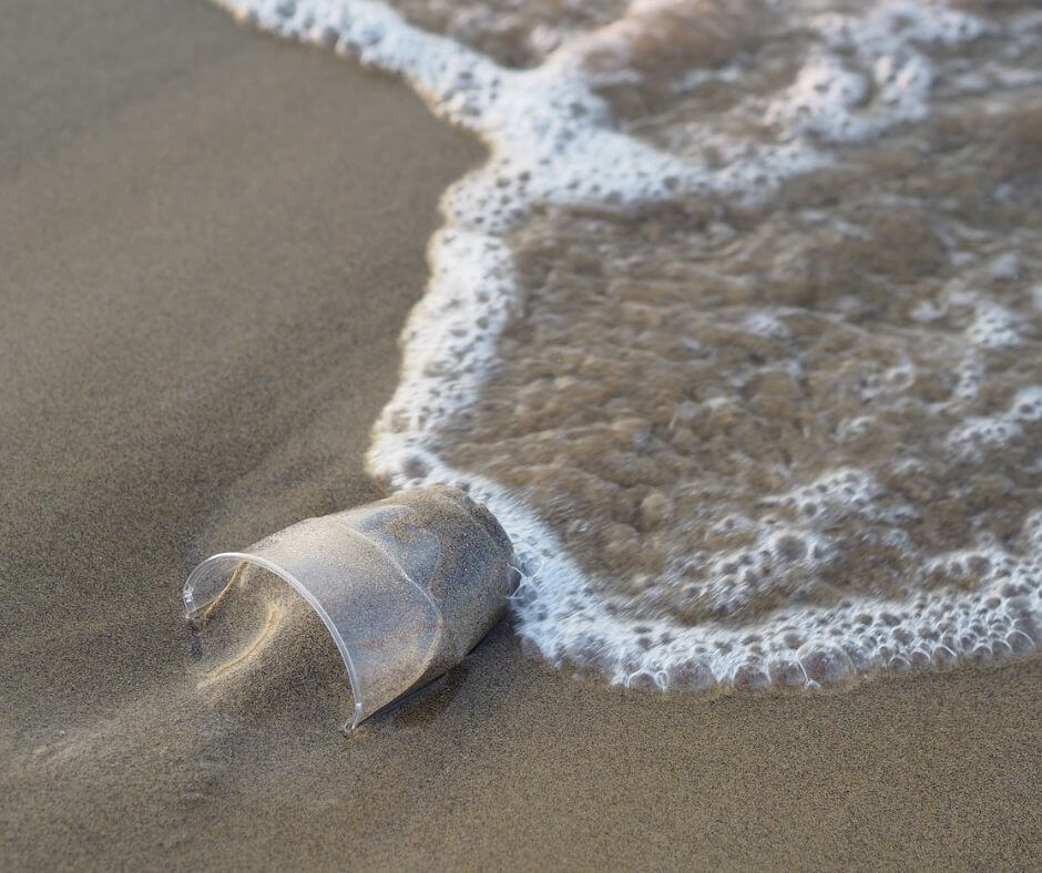 Ein Plastikbecher am Strand. Man sieht eine Welle, die bald den Plastikbecher erreicht
