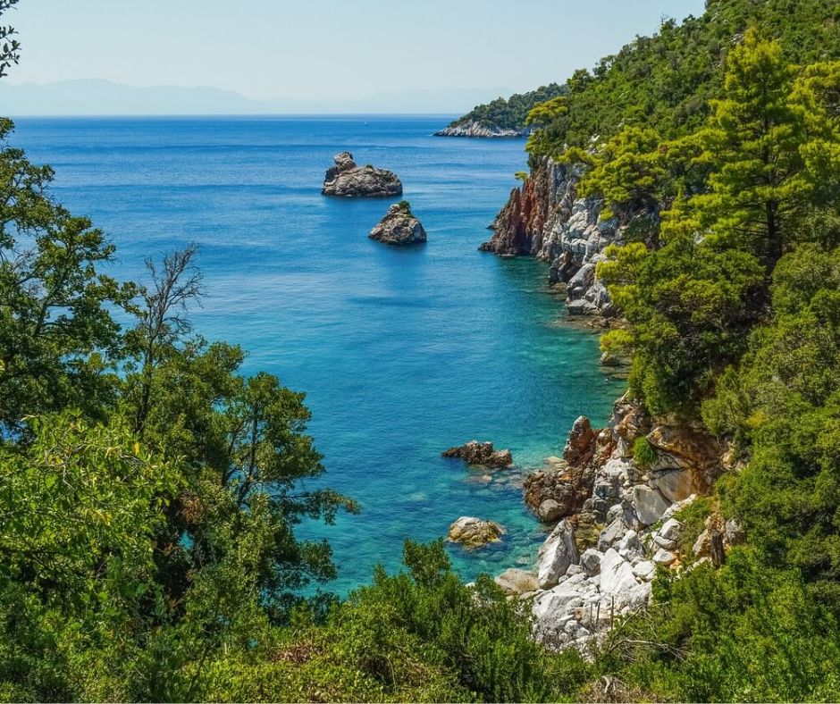 Begrünte Küste mit Felsen, die immer wieder aus dem Meer ragen. Im Zentrum das türkisfarbene Wasser. Im Hintergrund sieht man leichte Umrisse einer anderen Insel.