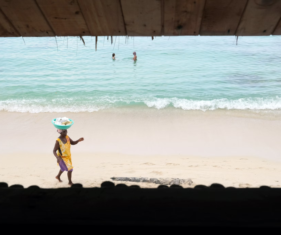 Una mujer caminando por la playa, llevando un cuenco encima de la cabeza. De fondo, 2 bañistas en una playa. 