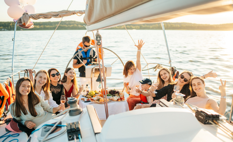 foto di un gruppo di amici su una barca a vela che fanno un picnic, dietro di loro uno skipper al timone