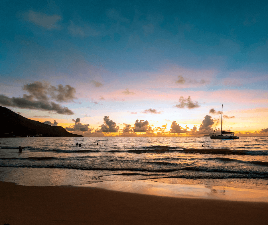 Catamaran anchored off a beach in Seychelles at sunset. People are bathing. 